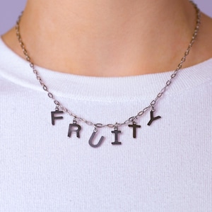 May include: A silver-toned metal chain necklace featuring individual letter charms that spell out the word FRUITY. The necklace consists of a delicate link chain with six distinct capital letter pendants hanging from the centre of the chain.