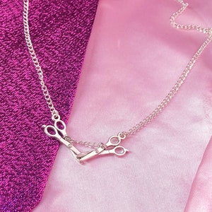 May include: A silver necklace with a scissors pendant. The pendant shows a detailed pair of scissors, linked by a fine chain. The necklace is displayed on a pink and purple fabric background.