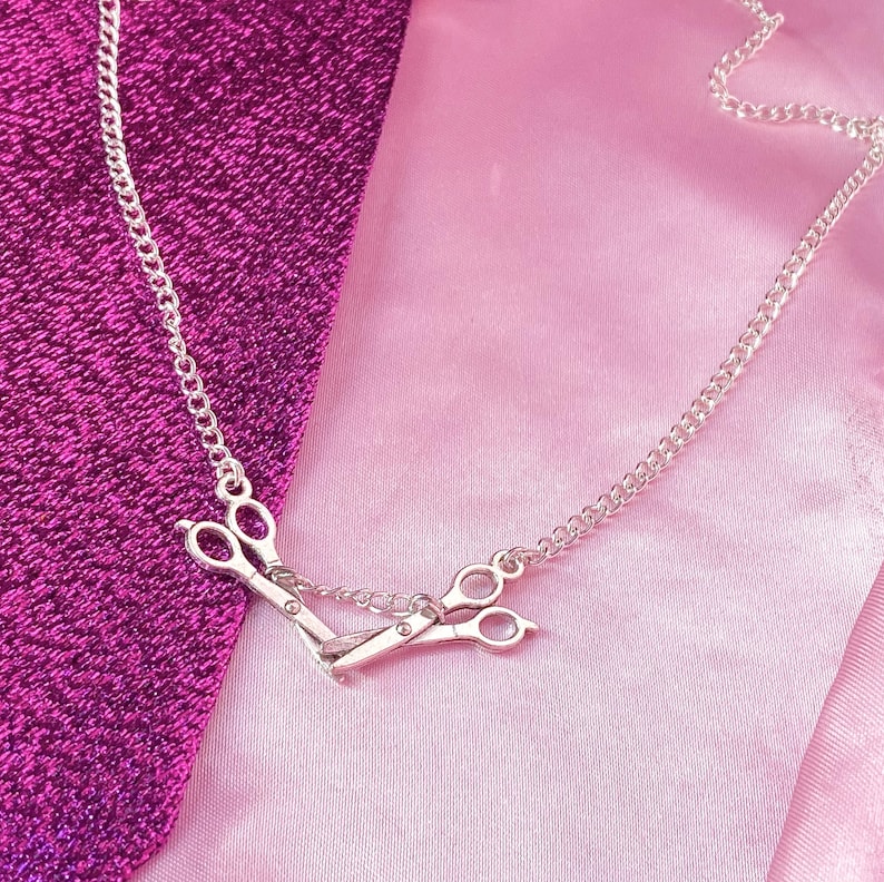May include: A silver necklace with a scissors pendant. The pendant shows a detailed pair of scissors, linked by a fine chain. The necklace is displayed on a pink and purple fabric background.