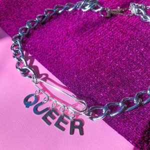 Peut inclure: Un collier en chaîne argentée avec un charme en forme d'épingle de sûreté et le mot "QUEER" écrit en charmes argentés. Le collier est sur un fond scintillant rose et violet.