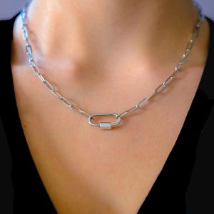Può includere: Una collana a catena color argento con chiusura ovale. La collana è composta da maglie a catena ovali. La chiusura è di forma rettangolare con una piccola barra. La collana è indossata intorno al collo.