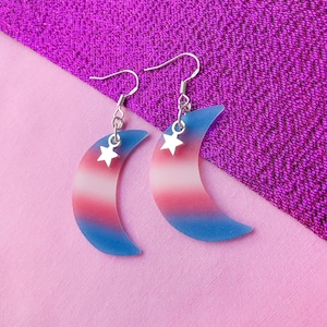 May include: A pair of crescent moon earrings with a gradient of pink, white, and blue. Each earring has a small silver star charm.
