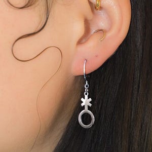 May include: A silver earring with a genderqueer symbol, hanging from a small hoop. The symbol is a combination of a circle with a cross above it and a line extending from the circle. The earring is worn on an ear with multiple piercings.