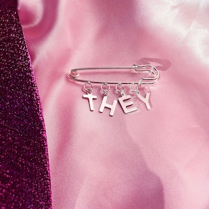 May include: A silver safety pin with five silver charms spelling out the word "THEY" hanging from it. The pin is on a pink satin fabric background.
