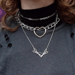 May include: A multi-layered silver necklace set. The top layer is a fine chain, the middle layer is a chunky chain with a heart pendant, and the bottom layer has a scissors pendant. The person is wearing a dark grey distressed t-shirt.