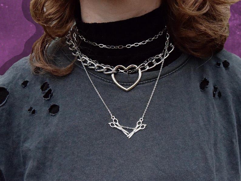 May include: A multi-layered silver necklace set. The top layer is a fine chain, the middle layer is a chunky chain with a heart pendant, and the bottom layer has a scissors pendant. The person is wearing a dark grey distressed t-shirt.