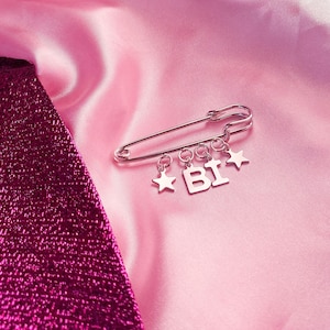 Könnte beinhalten: Silberne Sicherheitsnadel mit Anhängern, die "BI" und zwei Stern-Anhänger buchstabieren. Die Nadel wird auf einem rosa Satinhintergrund mit einem strukturierten, dunkelrosa Stoff in der Ecke präsentiert.