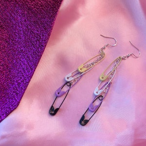 May include: A pair of dangle earrings featuring a chain with multiple safety pins in purple, yellow, and white.