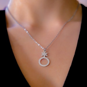 Op de afbeelding: Zilverkleurige ketting met een hanger met een genderqueer symbool. De hanger is een gestileerde combinatie van een cirkel, een kruis en een ster. De ketting wordt gedragen tegen een zwarte top.