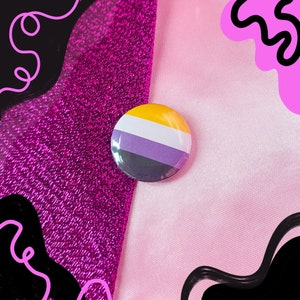 May include: A round button featuring the colours of the non-binary pride flag. The button is yellow, white, purple, and black. It is displayed against a pink and purple background.