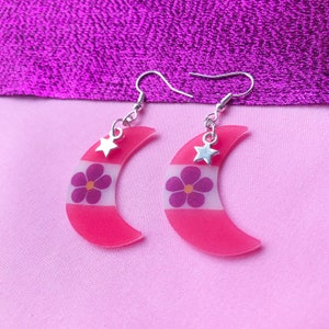 May include: A pair of pink crescent moon earrings with a white stripe and a purple flower design. Each earring has a small silver star charm.