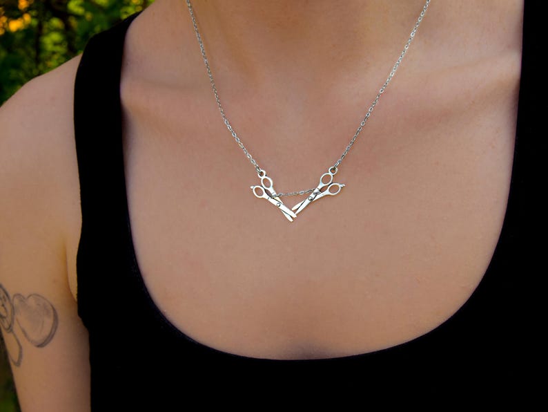 May include: A silver necklace with a pendant shaped like scissors. The scissors are joined at the handles and suspended from a fine chain. The necklace is worn over a black top.