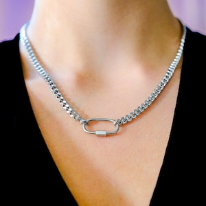 Peut inclure: Un collier en chaîne de couleur argentée avec un fermoir de type mousqueton. Le collier présente un motif de chaîne gourmette et se porte autour du cou. Le fermoir est de forme rectangulaire avec une petite barre.