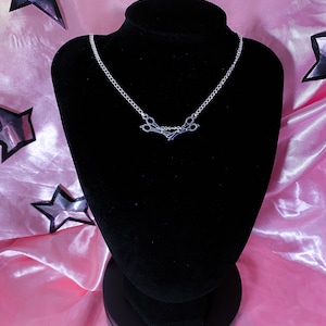 May include: A silver necklace with a scissors pendant, displayed on a black velvet bust. The background is pink satin, decorated with star shapes and fairy lights, creating a decorative display.
