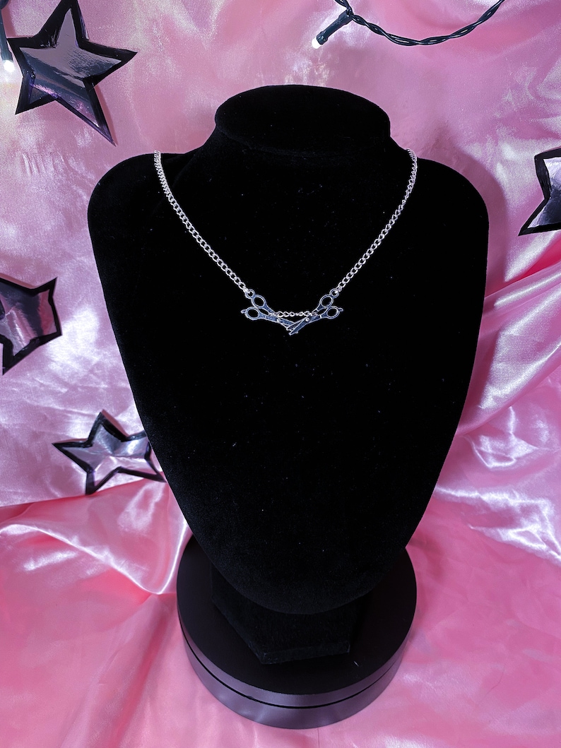 May include: A silver necklace with a scissors pendant, displayed on a black velvet bust. The background is pink satin, decorated with star shapes and fairy lights, creating a decorative display.