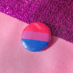 Può includere: Un bottone rotondo con una combinazione di colori rosa, viola e blu, che rappresenta la bandiera del pride bisessuale. Il bottone è posto su uno sfondo rosa e viola.