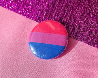 May include: A round button with a pink, purple, and blue color scheme, representing the bisexual pride flag. The button is set against a pink and purple background.
