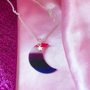 May include: A silver chain necklace with a crescent moon pendant. The moon is made of resin and has a gradient of red, white, black, and blue colours. A small silver star charm is attached to the top of the moon.