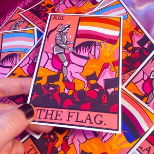 May include: A pink and orange tarot card with a white border. The card depicts a knight riding a unicorn, holding a flag with the colors of the lesbian pride flag. The card is titled "The Flag."