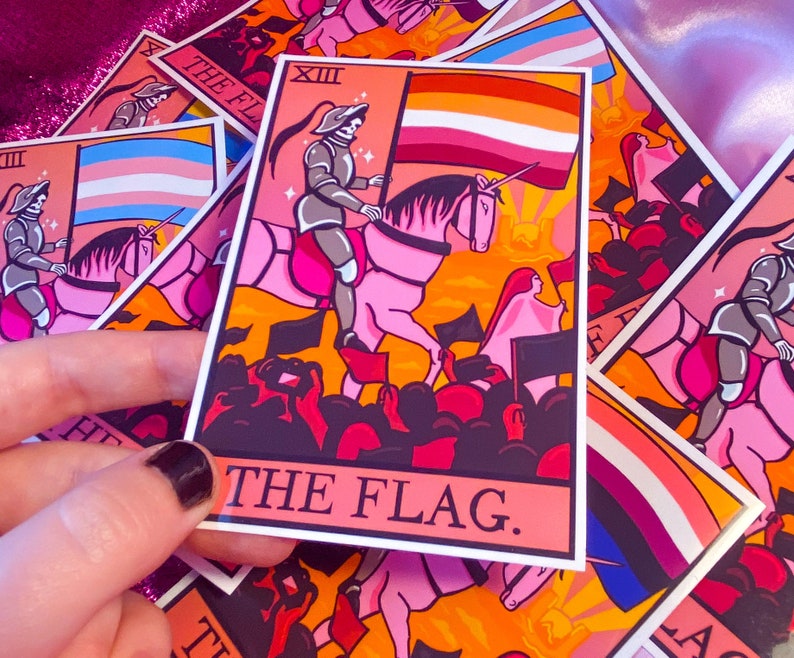 May include: A pink and orange tarot card with a white border. The card depicts a knight riding a unicorn, holding a flag with the colors of the lesbian pride flag. The card is titled "The Flag."