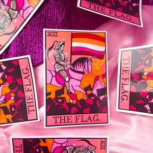 May include: A tarot card with a pink unicorn and a knight riding it. The card is set against a crowd of people and a sunset. The card is labeled "The Flag" and has the number "XIII" in the top left corner. The flag in the background is orange, white, and pink.