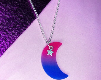 Puede incluir: Un collar de cadena plateada con un colgante de media luna. La luna está hecha de resina rosa y azul y tiene un pequeño amuleto de estrella plateada.