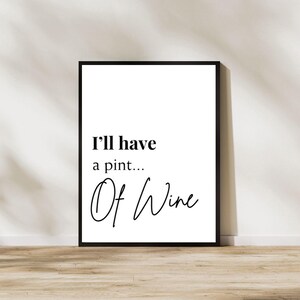 May include: A framed print with the text "I'll have a pint... Of Wine" in black script. The print is in a black frame and leans against a white wall. The background is a light wood surface.