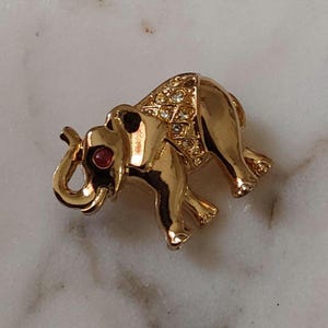 May include: Gold-toned elephant brooch with a red eye and a diamond-patterned back. The elephant's trunk is curled, and the brooch is set on a marble surface. A vintage-style jewelry piece.