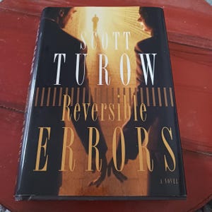 May include: A hardcover book titled Reversible Errors by Scott Turow. The black dust jacket features a silhouette of two people facing each other with a bright light in the background and a small figure walking toward the light in the distance.