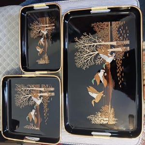 May include: Set of three black serving trays with gold trim and handles. Each tray features a decorative design of ducks in flight near trees. The trays vary in size, with the largest measuring approximately 18 inches long.
