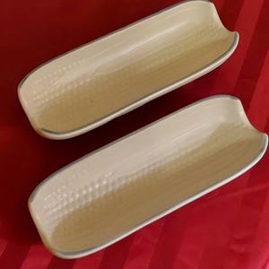 May include: Two elongated, cream-colored ceramic dishes designed in the shape of corn cobs. Each dish features a textured surface mimicking corn kernels and a smooth, curved interior with a light grey rim along the edges of the elongated form.