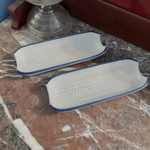 May include: Two white ceramic corn on the cob serving dishes with blue trim. The dishes are designed to hold corn on the cob.
