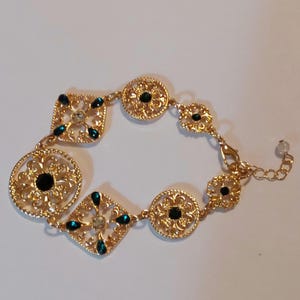 May include: Gold-toned bracelet featuring ornate filigree designs with square and circular elements. The bracelet is adorned with dark green and clear gemstones. The clasp has an extender chain with a small, round, light-colored bead.