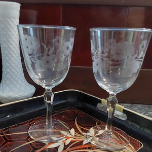 May include: Two clear glass wine glasses with etched floral designs on a black and gold tray.