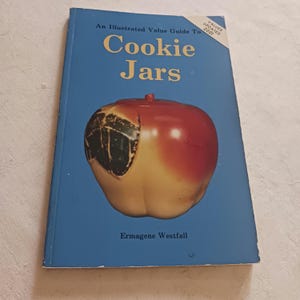 May include: A softcover book titled An Illustrated Value Guide To Cookie Jars by Ermagene Westfall. The blue cover features a large red and yellow apple-shaped cookie jar with a black leaf detail and a corner banner stating Values Updated 2001.