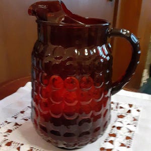 May include: A vintage ruby red glass pitcher with a textured, bubble-like pattern. The pitcher has a handle and a spout for pouring. The glass is a deep, translucent red, and the pitcher is sitting on a white lace doily.