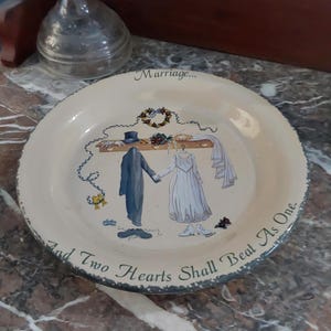 May include: A cream-colored decorative plate with a dark, distressed rim. The center features an illustration of a suit and wedding dress hanging on a rack, with the text "Marriage..." above and "And Two Hearts Shall Beat As One." below.
