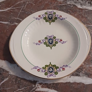 May include: White oval serving dish with a green and gold rim. The dish is decorated with a floral pattern in shades of purple, green, and red. The center of the dish features a repeating design of a blue and white crest with a floral motif.