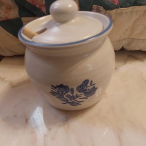 May include: A white ceramic sugar bowl with a rounded body and a matching lid featuring a small knob. The bowl is decorated with a blue floral pattern on the front and a thin blue rim around the lid. A small wooden spoon rests inside the bowl.
