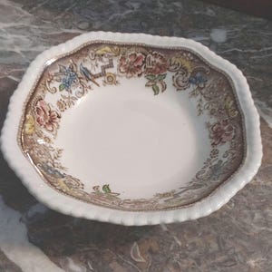 May include: A white, scalloped-edge bowl with a floral and bird pattern in shades of brown, pink, blue, and yellow. The rim of the bowl has a decorative, textured design. The bowl is likely made of ceramic or porcelain.