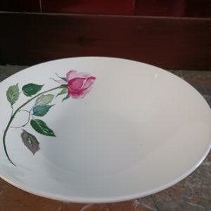 May include: A white ceramic plate with a pink rose and green leaves painted on the side. The rose is in full bloom, and the leaves are detailed. The plate is round and has a slightly raised edge. A vintage-style decorative plate.