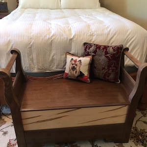 Peut inclure: Un banc de rangement en bois avec un dessus brun et un fond rayé blanc et brun. Le banc a deux accoudoirs et est décoré de deux coussins, l'un avec un motif de chien et l'autre avec un motif floral.