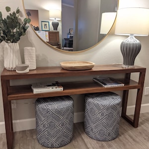 May include: A wooden console table with a shelf, a round mirror, two blue and white patterned ottomans, and a gray ceramic lamp with a white lampshade. The table has a wooden bowl and a book titled "Home" on the shelf.