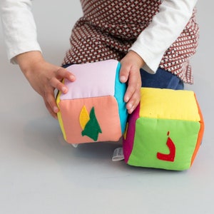 May include: Two colorful fabric blocks with different letters and designs. The blocks are pink, orange, yellow, green, and blue. The letters on the blocks are a green "A" and a red "J".