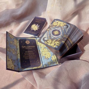 May include: A Spellbound Tarot Deck set featuring a purple box with gold floral accents, a matching guidebook, and a deck of cards. The box interior displays text including the title Spellbound Tarot Deck and a message to the reader. The card backs feature a central star, crescent moons, and yellow floral illustrations on a dark purple background.