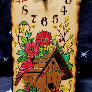 May include: Wooden clock with a rustic design. The clock face features black numerals and hands, with a red second hand. The clock is decorated with a birdhouse, red flowers, and red birds. The clock measures approximately 12 inches in height.