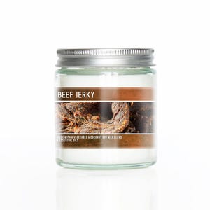 May include: A glass jar candle with a brown label that says "Beef Jerky" and "Made with a vegetable & coconut soy wax blend & essential oils". The label also features a close-up image of beef jerky.
