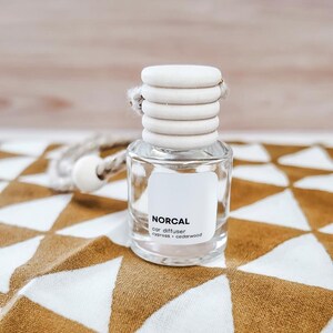 May include: A clear glass car diffuser bottle with a white label reading NORCAL car diffuser cypress + cedarwood. The bottle features a ribbed white wooden cap and a braided rope cord with a small white wooden bead attached for hanging.