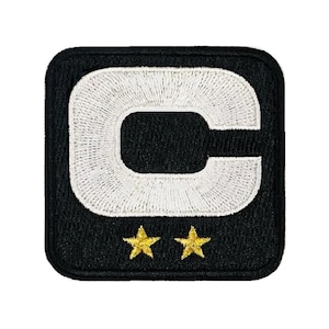 May include: A square-shaped embroidered patch with a black background featuring a large white letter C in the center. Below the letter C, there are two small, five-pointed gold stars positioned side by side. The patch has a black stitched border.