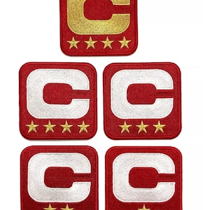 Könnte beinhalten: Eine Sammlung roter, quadratischer, gestickter Aufnäher. Jeder Aufnäher zeigt einen großen Buchstaben "C" in Gold oder Weiß, mit Sternen darunter. Die Aufnäher sind wahrscheinlich für Kleidung oder Accessoires.